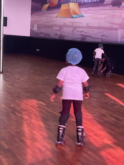 A person with a blue hair covering is wearing roller skates, knee pads, and wrist guards on a hardwood floor. Three people in similar safety gear are in the background. A painted wall is on the left. The location is not discernible as Moba, Nigeria.