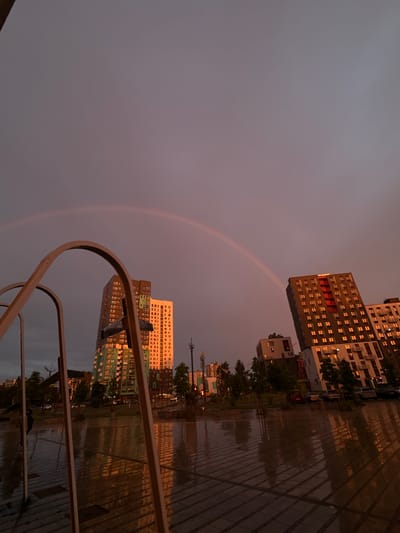 This image captures an urban or suburban scene in Бараўлянскі сельскі Савет, Belarus, at what appears to be dusk or pre-sunset, characterized by a predominantly grey-purple sky. A faint, diffuse double rainbow is discernible, arching across the upper portion of the sky, with its peak slightly left of the frame's center.
The scene is dominated by multi-story residential or commercial buildings. On the left, a tall, architecturally complex building with a dark facade and numerous windows reflects intense orange light from the low sun. Adjacent to it stands a lighter-toned, rectangular building, also displaying windows illuminated with an orange hue. On the right side of the frame, another prominent multi-story building, dark in color with red-accented balconies or architectural features, features numerous windows reflecting the same vivid orange light.
The foreground is occupied by large, curving, metallic structures, possibly arches of a monument or park feature, framing the lower left portion of the image. The ground is paved, wet, and highly reflective, mirroring the buildings and sky, indicating recent rainfall. Several small trees are planted in the mid-ground, their forms also reflected in the wet pavement. Faintly visible vehicles are present in the mid-ground between the trees and buildings. A partially obscured human figure is barely discernible through the arches on the far left side of the frame. The overall lighting suggests a warm, atmospheric glow from a setting sun reflecting off the clouds and wet surfaces.