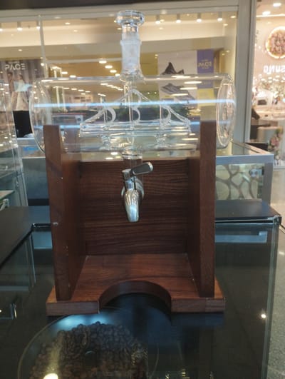 Here's a detailed description of the image:
**Subject:** The primary subject of the image is a glass beverage dispenser with a wooden stand. The dispenser is cylindrical with a narrow neck and stopper at the top and a metal spigot in the middle. Inside the glass container, there's a decorative glass element, possibly mimicking a chandelier or ornate structure. Beneath the dispenser, a clear glass plate holds coffee beans.
**Objects:**
* Glass Beverage Dispenser: The main focus.
* Wooden Stand: Holds the dispenser. Constructed with vertical panels on the side and solid panels on the bottom.
* Metal Spigot: Located in the middle of the dispenser.
* Glass Plate: Holds coffee beans.
* Coffee Beans: Dark roasted, scattered on the plate.
* Glass Cabinets: Partially visible on either side of the dispenser.
**Environment:** The environment appears to be a commercial setting, possibly a shop or a café/restaurant within a mall. The dispenser is sitting on a polished black surface, likely a countertop or display case. In the background, large windows showcase various retail stores. Banners and displays are visible through the windows, suggesting an indoor shopping area.
**People:** There are no people visible in the foreground. The image is focused solely on the object.
**Actions/Interactions:** No specific actions or interactions are depicted in the image. It seems to be a product display.
**Location:** The image does not provide concrete information to determine the specific location in Valencia, Venezuela. The commercial setting suggests it could be inside one of the malls or shopping centers in the city.
**Overall Impression:** The image portrays a static, well-lit product display in a commercial environment. The composition directs the viewer's attention to the beverage dispenser, showcasing its design and presentation.