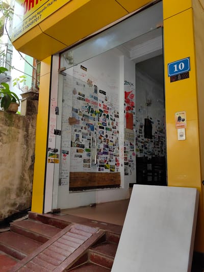 The image captures the entrance to a building in Thành phố Hà Giang, Vietnam. The building's address, "Số 10 - Đường Nguyễn Thái Học," is displayed on a yellow panel above the entrance, along with the MSDN number 5100497017. The entrance consists of a glass door surrounded by a yellow frame. The walls inside the entrance are densely covered with a variety of stickers, notes, and photographs. A blue sign with the number "10" is fixed to the yellow frame on the right side. Below it, there is another smaller sign. Further down on the right yellow frame is a "Xin Vui lòng BẤM CHUÔNG PLEASE RING BELL" sign above a bell button. The entrance is accessed via a set of red-brown brick steps that includes a ramp. A large white object partially blocks the lower right corner of the frame.