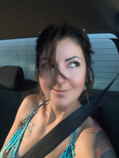 The image presents a head-and-torso view of a woman seated in the back seat of a vehicle. She has dark hair, styled away from her face, with some strands falling over her forehead and left eye. Her eyes are light brown, and she has dark, defined eyebrows. A metallic septum ring is visible on her nose. She is looking upwards and to her right with a slight smile. An earring, appearing to be a crescent moon charm with a chain, is visible on her left ear. She is wearing a teal or light blue patterned dress with a halter-neck or thin strap, which is partially visible on her right shoulder. A black seatbelt crosses her chest and left shoulder. A faint, light-colored tattoo is visible on her upper chest, partially obscured by the dress strap and seatbelt. A larger, dark tattoo with abstract shapes and red accents covers her left shoulder and upper arm. The background shows the dark interior of the car, including a headrest on the left, and a rear window with horizontal lines. Strong, warm light illuminates her face and right shoulder from the left side of the frame, creating highlights.
