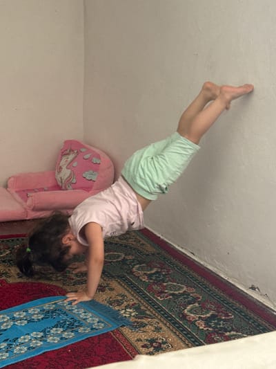 A young girl is captured in an upside-down yoga pose, seemingly balancing on her hands or forearms against a textured white wall. She wears a light pink t-shirt and light green shorts. Her legs are extended upwards, and her feet are pointed. Beside her, a large pink plush toy, shaped like a cushioned bed or seat, features a unicorn and clouds design. Below this, a patterned rug with intricate floral motifs in red, green, and gold is visible. A smaller blue rug with a white floral pattern lies on top of the larger rug. The lighting suggests an indoor setting, possibly during the daytime. The overall scene conveys a playful and active moment. The location context is Ləcət, Azerbaijan.