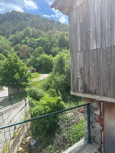An elevated, rotated landscape perspective reveals a wooden-planked structure at the upper left. Adjacent, there is a concrete element with stone and brick. A green metal fence spans part of the scene. The middle ground is dominated by verdant trees and vegetation. A road, a structure, and a light-colored vehicle are visible in the distance. A hillside forested with pine trees occupies the left horizon under a partly cloudy blue sky. The location appears to be Breza, Bulgaria.