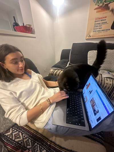 A woman is lying on a couch with a laptop on her lap, working online, while a black cat obstructs her view. The laptop screen displays a website in French titled "LA SOURC" and "LE GESTE". The couch has multiple pillows, and the woman is wearing a white shirt and several bracelets. In the background, there's a wall with a framed movie poster that says "UN FILM EN EASTMANCOLOR," which lists the actors and production team. The movie is a Franco-Italian production, CHAMPS ELYSEES PRODUCTIONS, PARIS. There's also a shelf with bottles and plates visible in the background.
