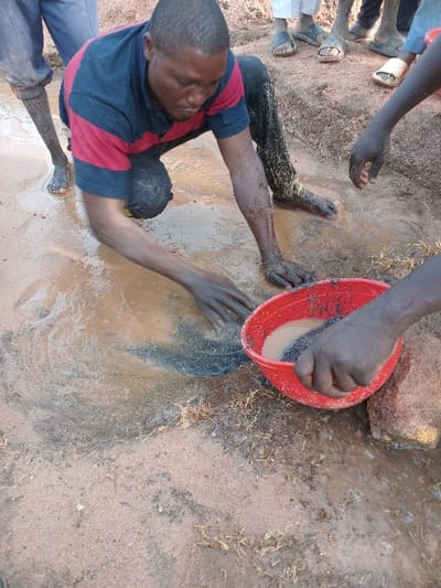 A man in a red and blue shirt kneels in mud, his arms outstretched and covered in dirt. Another person pours water from a red bowl onto the muddy surface. Multiple pairs of legs and feet, some wearing sandals, are visible around the scene. The ground is mostly reddish-brown dirt with patches of mud and some sparse vegetation. A rock is partially visible next to the bowl.