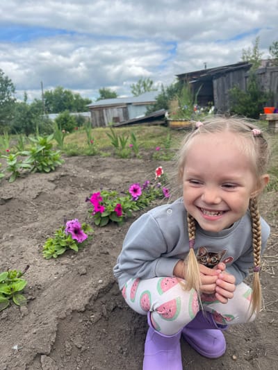 A young girl with blonde hair in two braids, wearing a grey sweatshirt with a cat graphic and white pants with a watermelon print, crouches in a garden. She is smiling broadly, with her eyes closed. In the background, there are several small buildings, trees, and a cloudy sky. The garden appears to be in a rural setting, likely in or near the village of Сухринское, Russia. The ground is dark soil, with some green plants and pink and purple flowers visible. The girl is wearing purple boots.