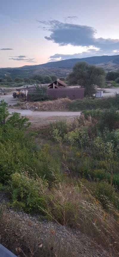 In Kocherinovo, Bulgaria, the image captures a rural landscape at dusk, featuring a cluster of buildings and natural elements. A yellow loader, a heavy construction vehicle, is parked near a covered structure, suggesting industrial or agricultural activity. Adjacent to it stands a building with a red, corrugated metal roof, appearing weathered and aged. Further to the right, a smaller, lighter-colored structure with a blue-tinted window and white trim is visible. The area is surrounded by abundant vegetation, including dense bushes, tall grasses, and scattered trees, with a large, mature tree dominating the mid-ground. In the background, rolling hills are silhouetted against a sky filled with soft, scattered clouds, illuminated by the fading light of sunset. The terrain appears uneven, with a gravelly ground surface around the structures.