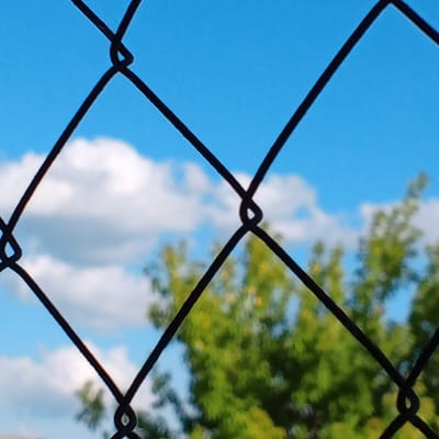 This is a close-up, eye-level, medium shot photograph taken outdoors during the daytime, looking through a black chain-link fence. The fence is a prominent foreground element, with its diamond-shaped openings creating a grid-like pattern across the frame. Beyond the fence, the background is composed of a bright blue sky with scattered white clouds. In the middle ground, there is a blurred green tree with some yellow foliage visible, suggesting it may be late summer or early autumn. The image does not contain any people or specific landmarks that would allow for identification of its location within the city of Khabarovsk, Russia. The focus is primarily on the textures and patterns of the fence against the natural background.