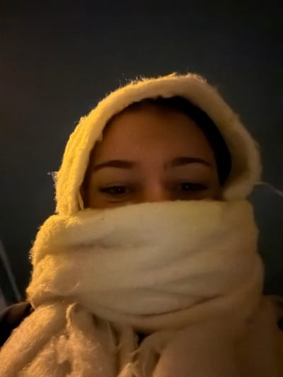 A person's face is seen from a low angle, partially obscured by a thick, cream-colored, textured scarf or hood. Their eyes and eyebrows are visible, with dark hair framed by the hood. The lighting is dim, casting shadows on the person's face and the fabric surrounding it. The background appears dark, possibly indicating an outdoor setting at night or in low-light conditions. The composition is intimate, focusing on the person's gaze.