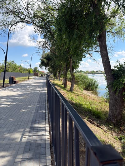 This image depicts a paved walkway in Leningradskaya, Russia, running alongside a body of water bordered by trees and vegetation. The walkway is constructed of light-colored rectangular pavers, with shadows cast across it from overhead trees. A dark metal railing runs along the edge of the walkway, separating it from a grassy embankment leading down to the water. Several streetlights are visible along the walkway, and a bench and trash receptacle are positioned on the left side of the path. The water is calm, reflecting the blue sky and scattered white clouds. On the far side of the water, buildings and more greenery are visible. The overall impression is that of a well-maintained public area, likely a park or promenade.