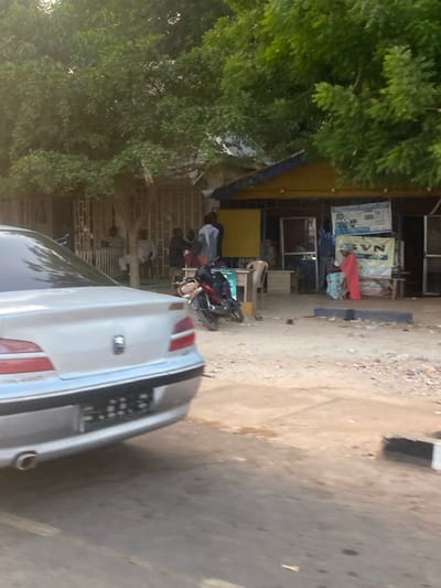 The image captures a detailed street-level scene in Birnin Kebbi, Nigeria. In the immediate foreground, the rear of a silver Peugeot 406 sedan is visible on the left, facing away from the viewer, with an indistinct license plate. The ground transitions from a paved or hard-packed surface to an unpaved, dusty area scattered with minor debris.
In the midground, a dark-colored motorcycle is parked on its kickstand in the dusty area. To the left, under a prominent canopy of green trees, several men are seated on chairs outside a building with horizontal slat-like window treatments; at least three individuals are discernible, one in light-colored attire, the others in darker clothing.
Further to the right, in front of a single-story building featuring a blue-and-yellow awning or fascia, multiple individuals are standing. Two men, one in a white garment and another in dark clothing, appear to be interacting. To the far right, a person dressed in a full-length, bright orange-red garment stands beside a wooden stand displaying a "BVN" sign and other advertisements. Various plastic chairs and small tables are positioned in this area. The background is dominated by the dense green foliage of several large trees, obscuring much of the buildings' roofs. The overall lighting suggests natural daylight, possibly late afternoon or early morning.