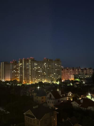 The image captures a nighttime aerial view of the city of Podkletnoe, Russia. In the background, a series of tall, illuminated apartment buildings dominate the skyline, their windows glowing with warm light. Red lights are visible atop some of these buildings. In the foreground, a dense cluster of houses with dark roofs and lit windows is nestled amongst trees, creating a stark contrast with the urban backdrop. Streetlights dot the landscape, casting pools of light onto the streets and rooftops. The sky is a deep, dark blue, with a few faint stars visible. The overall impression is one of a quiet, residential area at night, with the modern apartment blocks serving as a distant, glowing presence.