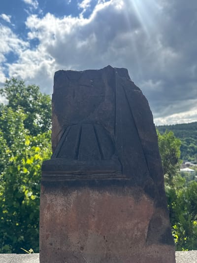This media file is an outdoor, eye-level shot featuring a weathered stone monument with intricate carvings. The monument is dark in color and appears to be made of rough-hewn stone, possibly basalt, with areas of lighter, reddish-brown discoloration indicating age and exposure. The carvings on the monument depict geometric patterns and a stylized, fan-like motif at the top, suggesting it might be part of an ancient structure or marker.

The background of the image is dominated by a bright, partly cloudy sky. Sunlight streams through the clouds, creating a dramatic effect with visible light rays. Lush green trees with dense foliage occupy the lower portion of the frame and extend into the middle ground, suggesting a natural or park-like setting. In the distance, through the trees, hints of buildings or structures can be seen, along with a hillside covered in vegetation.

The overall impression is one of antiquity and nature. The lighting suggests it is daytime, likely mid-morning or afternoon, given the bright sun and clear blue sky patches visible through the clouds. The weather appears to be fair with scattered clouds. The combination of the ancient stone, abundant greenery, and distant structures points to a location with historical significance, possibly an archaeological site or a scenic historical area. The image does not contain any visible text. The setting is identified as Dilijan, Armenia.