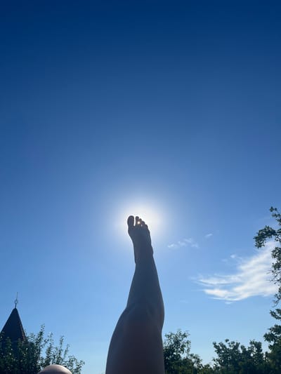 The image captures a unique perspective looking up at a clear blue sky with the sun as a prominent feature. In the foreground, a person's foot is silhouetted against the bright sun, with toes clearly visible. The leg and a portion of the arm are also visible, suggesting the person is lying down or extending their limbs upwards. The scene is outdoors, with dappled sunlight filtering through lush green foliage visible at the top and right edges of the frame, hinting at trees overhead. In the lower right corner, a dark, angular structure, possibly a roof or building, is partially visible, adorned with a small, metallic finial. Wispy white clouds are scattered across the sky, adding texture to the vast expanse of blue. The bright sunlight suggests it is daytime, likely a sunny afternoon, and the clear skies and greenery evoke a feeling of warmth and relaxation. The photograph seems to be taken in Stăuceni, Moldova, given the location context. There is no visible text in the image.