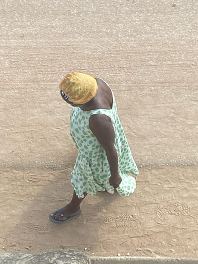 A high-angle, overhead image depicts a single individual walking on a light brown, unpaved surface in Ngleshi Amanfro, Ghana. The person, observed from behind and slightly to their left, has dark skin. They are wearing a sleeveless, light green dress with irregular dark green polka dots and a yellow, woven cap covering their head. Blue flip-flops are on their feet, with a small white object visible on the second toe of the left foot. The individual's head is tilted downward and to their right, and their left arm is bent. The ground is dry earth, marked with numerous faint shoe prints and tire tracks. A portion of a rough, light-colored concrete or stone curb is visible in the lower left foreground, delineating the edge of the walking surface.