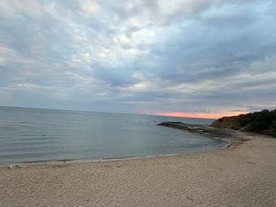 The image depicts a tranquil beach scene in Lozenets, Bulgaria, likely during a sunset or sunrise due to the soft, diffused light and the warm hues on the horizon. The foreground is a sandy beach, with visible footprints and the texture of the sand. In the mid-ground, the calm sea stretches out, with gentle waves lapping at the shore. A small, red buoy is visible in the water. Further in the distance, a rocky outcrop juts into the sea, with several small figures standing on it, possibly engaged in fishing or enjoying the view. To the right, a grassy, tree-lined cliff forms the edge of the coastline. The sky above is filled with dramatic, layered clouds, in shades of grey, white, and hints of pink and orange near the horizon. The overall atmosphere is one of peacefulness and natural beauty.