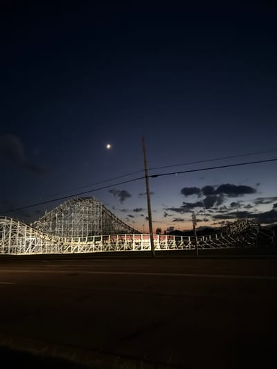 This is a nighttime photograph of a wooden roller coaster in Wisconsin Dells, USA. The image is oriented vertically, with the sky occupying the left side and the roller coaster on the right. The sky is a deep, dark blue, transitioning to lighter shades of gray and hints of orange near the horizon, indicating either twilight or dawn. Clouds are scattered across the sky, some appearing dark and dramatic, while others are illuminated by unseen light sources. A crescent moon is visible in the upper left quadrant of the sky.

The roller coaster is a large, intricate wooden structure with numerous tracks and supports, illuminated by bright artificial lights that highlight its framework. The design suggests a significant thrill ride. The roller coaster takes up a large portion of the right side of the frame. To the right of the roller coaster, a dark, solid mass likely represents the ground or a structure, with some faint details suggesting a road or pathway.

The scene conveys a sense of quiet anticipation or closure, as the roller coaster appears to be inactive. The time of day is clearly night, with the moon and fading twilight suggesting it's either late evening or early morning. The weather appears clear enough for celestial observation, with dramatic cloud formations adding to the atmosphere. There are no people visible in the image. The presence of the roller coaster and the overall setting strongly suggest an amusement park or theme park. No discernible text is visible in the image.