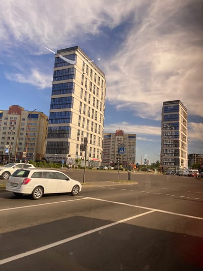 The image, taken in Brest, Belarus, shows an urban street scene with modern buildings and a road. The orientation of the image is rotated 90 degrees counter-clockwise, presenting a vertical perspective.
In the foreground, a white car is parked on the right side of the road, partially obscuring another car behind it. The road itself has white lane markings.
Dominating the background are several multi-story buildings. The largest building, a pale beige structure with multiple rows of windows framed by darker blue glass, is in the mid-ground. To its right, a yellow and blue apartment building is visible. Further in the distance, a taller, narrower beige building with similar window patterns stands out.
The sky is a bright blue, with wispy white clouds scattered across it, suggesting a clear, sunny day. The lighting indicates it is likely daytime, possibly late afternoon given the angle of the shadows.
Several street signs are visible, including one with a blue pedestrian symbol and another with a white "P" on a blue background, indicating parking. Some people can be seen walking on the sidewalk in front of the buildings.
A sign with Cyrillic text is visible on one of the buildings, though it is not legible enough to read. The overall scene depicts a typical urban environment with residential and commercial structures, a road, and vehicles.
