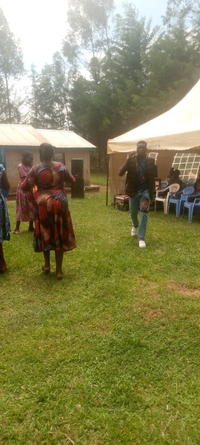The image shows a scene in a grassy outdoor area, likely a rural setting.  Several people are visible. Two women are dressed in brightly colored dresses with floral patterns. One woman is facing the camera, and the other is walking away from the camera.  A man, dressed in a brown jacket, blue jeans with a tear in the knee, and white sneakers, is walking toward the camera. He appears to be speaking on a mobile phone. In the background, a large, white tent is partially visible. Several blue chairs are situated around the tent.  The image was taken in Kivaywa, Kenya, at the coordinates (latitude: 0.56, longitude: 34.806).