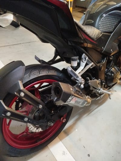This media file displays a close-up, rear-view of a black and red sport motorcycle parked indoors, possibly in a garage or parking lot. The motorcycle is the sole object of focus, with its components such as the rear tire, red rim, exhaust system, and part of the frame prominently featured.

The exhaust pipe has a carbon fiber finish and a metallic Yoshimura logo with "U.S.A." visible on it. The rear wheel is red, contrasting with the black tire. The scene is illuminated by artificial light, suggesting it is likely daytime or evening. There are no people visible in the frame. The ground appears to be concrete, and some smudges or dirt are present on it, indicating it's a functional space rather than a pristine showroom. The overall impression is a detailed look at a parked motorcycle, highlighting its performance-oriented features. The context of Phuket City Municipality, Thailand, does not provide specific visual cues within the image itself.