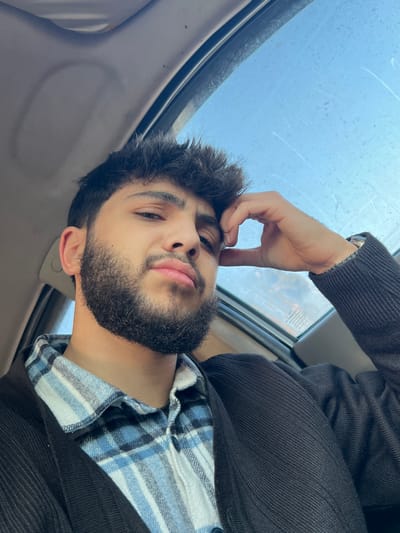 A young man with a beard poses in what appears to be a car interior. He wears a black jacket over a blue and white checkered shirt. His hand is near his forehead, possibly resting or posing. A window is visible in the background, seemingly showing a cloudy or rainy sky. The photo is taken at an angle, adding a dynamic element to the composition.