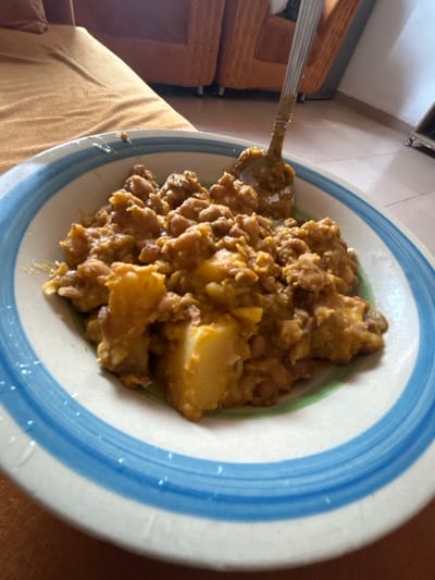 A bowl of food sits on a table. The bowl has a blue and white striped pattern. The food appears to be a mix of beans and potatoes, covered in a brown sauce or gravy. A spoon is resting inside the bowl, holding a small portion of the food. The food is rustic and looks homemade, suggesting a hearty, comforting meal.
