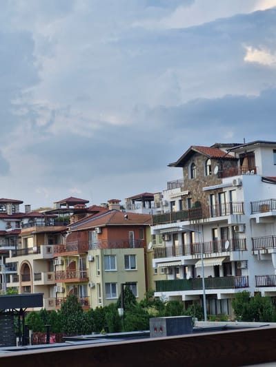 The image depicts a row of multi-story buildings with various architectural styles, predominantly featuring red tile roofs and white or light-colored facades. Many of the buildings have balconies with dark railings, and some are adorned with awnings. The buildings are situated on a slope, with some appearing to be hotels or apartment complexes. Trees and greenery are visible in front of and between the buildings. The sky is overcast with light-colored clouds. A small bird is visible in the sky. The overall scene suggests a residential or tourist area in a coastal town. This is a view of buildings located in the city of Sveti Vlas, Bulgaria.
