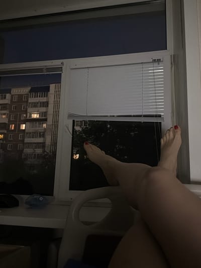 The image shows a close-up view of a person's legs and feet propped up against a window, with a view of a multi-story apartment building in the background. The scene appears to be indoors, possibly in an apartment or hotel room, with blinds partially covering the window. The lighting suggests it is nighttime or dusk. The apartment building in the background is typical of Soviet-era architecture, common in cities like Tomsk, Russia. The building has multiple floors with numerous windows, some of which are illuminated, indicating activity within the apartments. The presence of blinds suggests an attempt to control light or privacy. The overall atmosphere is quiet and domestic.