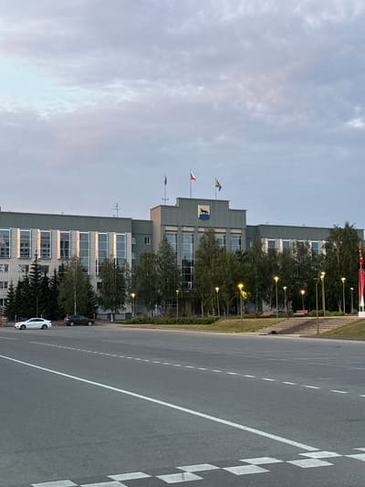 The image shows a modern, multi-story building with many windows, likely an office building or administrative center, in Surgut, Russia. The building is a light gray color and has a stepped architectural design. It is surrounded by trees and has flags flying on poles in front of it, including what appears to be a blue and yellow flag and a red and white flag. A crest with a black animal on a yellow background is also visible on the building.
In front of the building is a paved area, possibly a parking lot or street, with white lane markings. Two cars are parked on this paved area: a dark-colored car and a white car. The scene is well-lit, suggesting it is daytime, likely late afternoon or early evening, given the soft light and the presence of streetlights that are on. The sky is overcast with scattered clouds. The overall impression is one of an organized and functional urban environment. There is no visible text in the image.