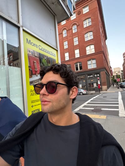 A male individual with dark, curly hair and red-tinted sunglasses is positioned in the foreground, looking to the right. He wears a dark t-shirt and a dark sweater draped over his shoulders, with a thin silver chain visible around his neck. To his immediate left is a storefront window displaying a yellow poster with the text "More Connection," "Art for Boston," and "BOSTON," featuring an artistic image.
In the midground, a prominent multi-story red brick building with arched windows on its upper floors stands on the right. Its ground level features a storefront displaying Chinese characters, including "波士頓" (Boston) and "華埠" (Chinatown), indicative of a business in Boston's Chinatown. A street corner is visible with a white striped crosswalk and yellow tactile paving. A silver sedan is parked on the street, and a red postbox and a dark utility box are on the sidewalk. Additional buildings and distant pedestrians are visible under a clear, light blue sky.
The scene is located at a street corner within Boston's Chinatown, United States.