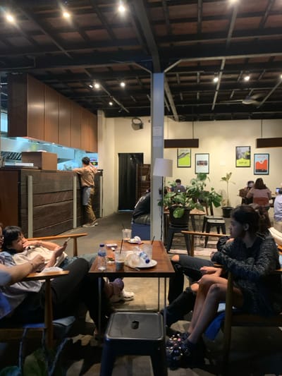 The image shows the interior of a cafe in Malang, Indonesia, located at coordinates -7.946 latitude, 112.626 longitude. There are three people seated at a table in the cafe, along with a man standing near a counter in the background. The cafe has a wooden ceiling with exposed beams. The walls are painted white and decorated with framed artwork. The table is made of wood and is surrounded by chairs. On the table are several empty glasses, a bottle of water, and a plate with some food. One person is sitting on a couch and looking at a phone. Two people are sitting across from each other, with the person on the right looking directly at the camera. A black stool is positioned in the center of the scene. The cafe has a modern and minimalist aesthetic. There are plants in the background, along with a partially visible counter. The lighting in the cafe is warm and inviting.