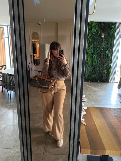 A woman is seen in a full-body reflection in a mirror. She is wearing a brown button-up shirt and beige pants and has a brown handbag hanging from her shoulder. She is holding up a phone, presumably taking a photo of herself in the mirror. The background reflected in the mirror shows a room with a table and chairs, a doorway, and a wall with some blue writing on it. The mirror is in a space that has a tiled floor, a green wall, and a wooden tabletop. Based on the interior design elements, it is possible that this image was taken in a cafe or restaurant. It is not possible to confirm the specific location in Marneuli, Georgia without further information.