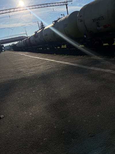 A train of tank cars is parked on railroad tracks next to a paved area with a white line down the middle. The sun shines brightly from the upper left corner of the frame, casting a lens flare across the image and creating shadows on the pavement. In the background, there is a clear blue sky with some clouds and overhead power lines. A pedestrian bridge spans the tracks in the distance. The tank cars appear to be empty or are carrying a liquid cargo, as indicated by their cylindrical shape and the markings on their sides. The scene suggests a transportation hub or industrial area in Kalynivka, Ukraine, likely during daylight hours.