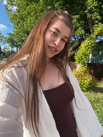 This is a vertically oriented, close-up selfie of a young woman with long, light brown hair, taken outdoors. She is smiling and looking towards the camera. She is wearing a dark brown top and a white, sheer garment draped over her shoulders and chest, possibly a scarf or light jacket. Her hair is long and flowing, some strands fall across her face and shoulders.

The background shows a bright, sunny scene with lush green trees and foliage. In the upper right portion of the image, part of a building with a blue-trimmed window is visible, suggesting a residential area. The sky above is a clear blue with a few white clouds. The lighting indicates it is daytime, likely late morning or afternoon, with strong sunlight casting shadows and highlighting the textures of her clothing and hair. The overall mood appears relaxed and cheerful.

The image was taken in or around Kurovskoye, Russia, indicated by the location context. No visible text is present in the image.