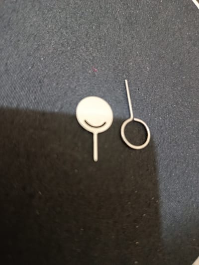 The image shows two SIM card ejector tools on a dark, textured surface, likely a fabric mat or cloth. One tool has a round, white head with a simple curved line etched into it, resembling a smiley face. The other tool is made of silver metal and features a loop at one end. The tools are small and appear to be for electronic devices like smartphones. The background is out of focus, but the texture suggests a woven material with some light-colored specks. There is no visible text in the image. The lighting appears to be artificial, casting a subtle shadow from the tools onto the surface. The scene is simple and focuses on the objects themselves.