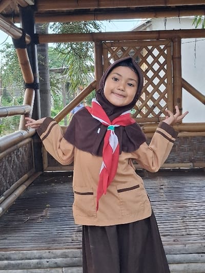 A young female subject is depicted standing centrally within an open bamboo structure in Kalitengah, Indonesia. She is wearing a dark brown hijab, a dark brown cape, a light brown long-sleeved uniform shirt with two dark brown stripes on each cuff and above two front pockets, and a dark brown ankle-length skirt. A red and white striped neckerchief is tied around her neck beneath the cape. The subject is smiling directly forward with a slight head tilt. Both arms are raised to approximately shoulder height, with elbows bent and hands open, fingers splayed. The immediate environment consists of a floor and structural elements made of horizontal bamboo slats and vertical bamboo poles, with a section of lattice-patterned bamboo visible in the background. Beyond the structure, green foliage, trees, and a segment of a light-colored building are visible under a bright sky, indicating an outdoor daytime setting.