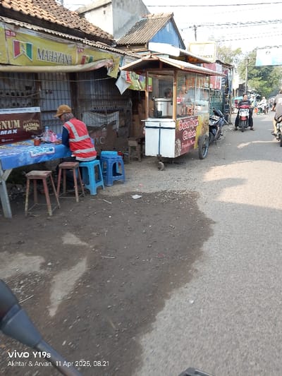 The image shows a street scene in Kecamatan Haurgeulis, Indonesia. A man wearing an orange vest is sitting at a small, blue and white table. He is seated on a blue stool, and two red stools are near him. The table is covered with a blue tablecloth. There is a food cart in the background. There are other people in the background, but they are not in focus. The street is paved and there is a small amount of dirt on the left side. A motorcycle is parked in the background. The sky is bright.