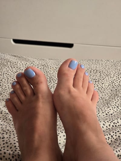 The image displays a close-up, overhead view of a person's feet, adorned with light blue nail polish. The feet are positioned on a surface covered with a black and white speckled fabric. In the background, the lower portion of a light-colored piece of furniture, possibly a dresser, is visible. The furniture has a horizontal dark line running across its front, suggesting a drawer or a recessed handle. The setting appears to be indoors, in a bedroom or living area. The image does not contain any information that would allow for localization to Petropavlivska Borshchahivka, Ukraine.