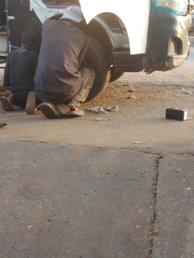 Two individuals are working on the front tire of a white van on a paved surface. One person is kneeling while the other is crouched, both focused on the tire. Tools and small parts are scattered on the ground nearby. A rectangular object sits slightly apart from the tools. The scene appears to be outdoors in daylight, possibly on a road or at a repair location.