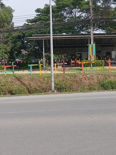 The image shows a roadside view of a building in Sakhu, Thailand, likely a restaurant or cafe, with an outdoor seating area. A paved road occupies the foreground, with a grassy embankment separating it from the building. The building has a corrugated metal roof supported by pillars, and appears to be open-fronted. Colorful wooden posts and some potted plants are visible near the building. Clothes pegs with small colorful ornaments are strung along a wire in front of the building. A utility pole with numerous wires runs vertically through the scene. The overall impression is of a casual, possibly commercial establishment located in a tropical environment, given the dense foliage and trees.