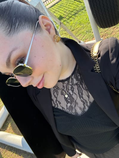 A person, likely a woman, is wearing a black jacket over a black top with lace detailing. She sports vintage sunglasses with gold frames and a chain embellishment on the jacket. A gold chain necklace and earrings add to the aesthetic. The photo is taken from a close angle, highlighting the person's face and upper body, with an outdoor background slightly blurred.