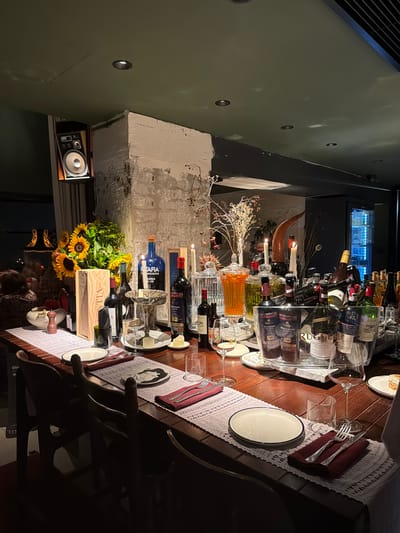The image captures a dimly lit, rustic-themed table setting, likely for a celebratory dinner or gathering. The table is laden with numerous wine bottles, some displayed in a clear acrylic rack. Various glasses, plates, and cutlery are arranged on white lace placemats. A centerpiece features sunflowers and dried floral arrangements, along with decorative candles and a unique horn-shaped object. There's also a decanter filled with an orange liquid.
In the background, a glimpse of a refrigerated unit with blue lighting suggests a bar or kitchen area. A vintage speaker system is visible on a shelf to the left. The walls appear to be a textured, light-colored material, possibly brick or plaster, with some dark structural elements. The overall atmosphere is intimate and dimly lit, creating a cozy and sophisticated ambiance. The arrangement suggests the preparation for a meal or a pause during one.
Visible text includes "VILNA" on a wooden wine crate and labels on several wine bottles, though the text on the bottles is mostly illegible due to the image quality and angle. The setting seems to be indoors, possibly a restaurant or a home dining area in Limassol, Cyprus, given the location context. The time of day is likely evening, given the artificial lighting and the overall mood. No people are visible in the frame.