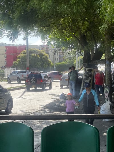 The image depicts a street scene in the Perímetro Urbano Barranquilla, Colombia. The orientation of the photo is sideways. In the foreground, two green plastic seats are visible, suggesting an outdoor seating area or public transportation stop. A dark metal pole runs vertically along the right side of the image, partially obscuring the view.
In the middle ground, several people are gathered under a large tree and a makeshift shelter. One man in a red shirt and jeans stands, while another in a blue and black jacket and a blue baseball cap appears to be talking to a woman in a pink top and orange hat. A third person, wearing a green shirt and jeans, is partially visible, standing near a light-colored car. Two other cars, a dark-colored SUV and a red vehicle, are parked along the street.
The background shows buildings with red and yellow facades, indicative of urban architecture in the region. Overhead power lines are strung across the scene. The lighting suggests it is daytime, likely late morning or early afternoon, with sunlight filtering through the leaves of the tree, casting shadows on the ground. The overall atmosphere appears casual, with people interacting outdoors. No explicit text is clearly legible in the image.
