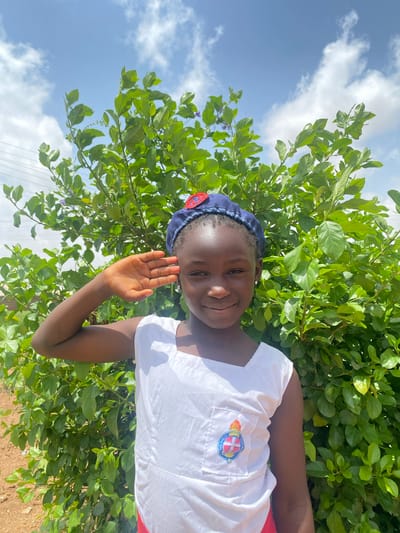 A young Black girl is standing outside in Turu, Nigeria. She is wearing a white shirt with a patch that says "The Girls Brigade" and a blue hat. She is smiling and waving her hand towards the camera. Behind her, there is a green leafy shrub and a blue sky with white clouds. The ground appears to be dirt.