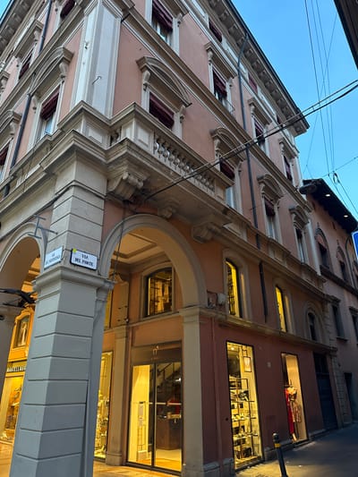 The image depicts the exterior of a building located at the intersection of Via Del Monte and Via Dell'Indipendenza in Bologna, Italy. The building is characterized by its pastel pink facade and classical architectural details, including arched entrances, balconies with balustrades, and decorative cornices.
Street signs clearly label the intersecting streets. The ground floor features shopfronts with large glass windows, showcasing merchandise. The shop on the left has a sign that reads "MANTELLASS." The shop on the right has a sign that reads "o'dett." The lighting from the shop windows suggests it is evening or nighttime.
Above the ground floor, windows are visible, some with red awnings. Overhead, power lines crisscross against the twilight sky. The overall impression is of a well-maintained historic urban environment. No people are explicitly visible in the image, but the presence of shops indicates commercial activity. The scene is static and appears to be a street-level view taken from a slightly upward angle.