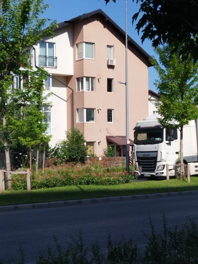 The image depicts a white DAF semi-trailer truck parked on the side of a paved road. The truck is facing towards the viewer, with its headlights illuminated. The road appears to be in an urban or suburban area, with a sidewalk and a grassy verge running alongside it. Behind the truck and verge, there is a modern building with a light beige and white facade, featuring multiple windows and balconies. Lush green trees and a flowerbed with red and pink flowers are visible in front of the building, suggesting a well-maintained outdoor space. The sky is clear and blue. The scene is static, with no people or apparent activity. This image was taken in Bucharest, Romania.
