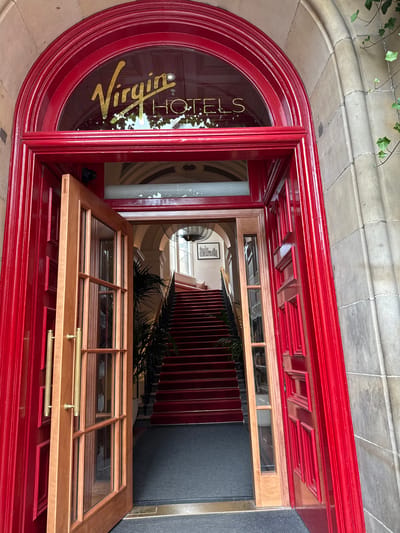A full exterior shot captures the grand entrance of Virgin Hotels Edinburgh. The facade features a large, arched red doorway with ornate trim. Above the doorway, a semicircular glass pane displays the hotel's name in gold lettering: "Virgin HOTELS". The main door is ajar, revealing a glimpse of the interior. To the left, a double wooden door with glass panes and brass handles stands open, offering a view of a carpeted staircase. The staircase, lined with red carpet and brass accents, ascends into the building. A large potted plant is visible to the left of the stairs. The surrounding architecture is of stone masonry.