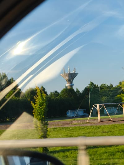 The image captures a view from inside a vehicle looking out a window, likely a car, at a landscape under a bright, sunny sky with streaks of clouds. In the distance, a tall, modern water tower with a distinctive flared top and antennae is visible, surrounded by lush green trees. To the right of the water tower, a section of a sports field is apparent, featuring a goal frame with a net and a bench structure. The foreground shows a reflection on the window, obscuring parts of the scene. The overall setting appears to be a park or an open area with greenery and sports facilities, likely within or on the outskirts of the city of Grobiņa, Latvia.