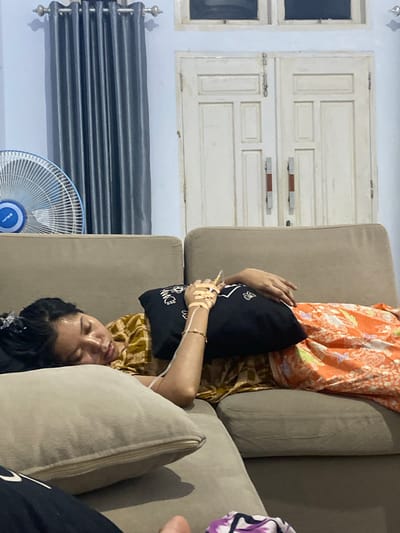 A woman lies on a brown couch, her head propped on a black pillow. She wears an orange shirt with a floral pattern and a brown and yellow checkered skirt. Her right arm is bandaged and connected to an IV drip. She appears to be sleeping or resting, with her eyes closed and her mouth slightly open.

In the background, a closed white door with two panes is visible, along with a window to its left. To the woman's left, a dark gray pleated curtain hangs above a white electric fan with a blue center. The scene appears to be indoors, possibly a living room. The lighting suggests it is evening or night. The presence of the IV drip indicates the woman may be unwell or recovering. The image is oriented vertically.
