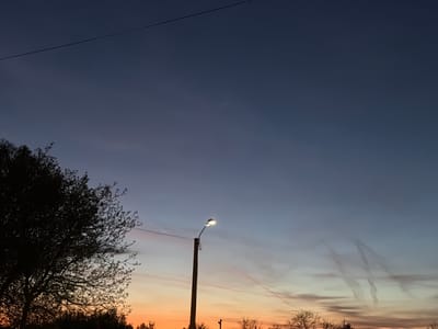 The image depicts an outdoor evening scene in Târgu Mureș, Romania, as suggested by the environmental context of the image.
**Environment:** The sky transitions from orange at the horizon to a deep blue further up. Contrails are visible in the sky. Trees with bare branches are silhouetted against the horizon.
**Objects:** A single lit streetlight is a central element, standing on a pole, connected to wires. An electrical cable stretches horizontally across the upper part of the frame. A tree appears on the left side of the frame.
**People/Subjects:** No people or animals are visible.
**Actions/Interactions:** The streetlight is illuminated. The sunset provides the backdrop.